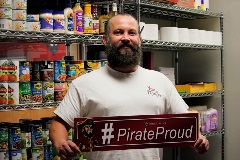 Thomas holds a #PirateProud sign inside of the Pirate Pantry with canned goods behind him